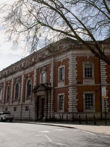 A historic brick building with large arched windows and a decorative entrance is surrounded by leafless trees. A parked vehicle is visible on the street in front, under a clear sky.