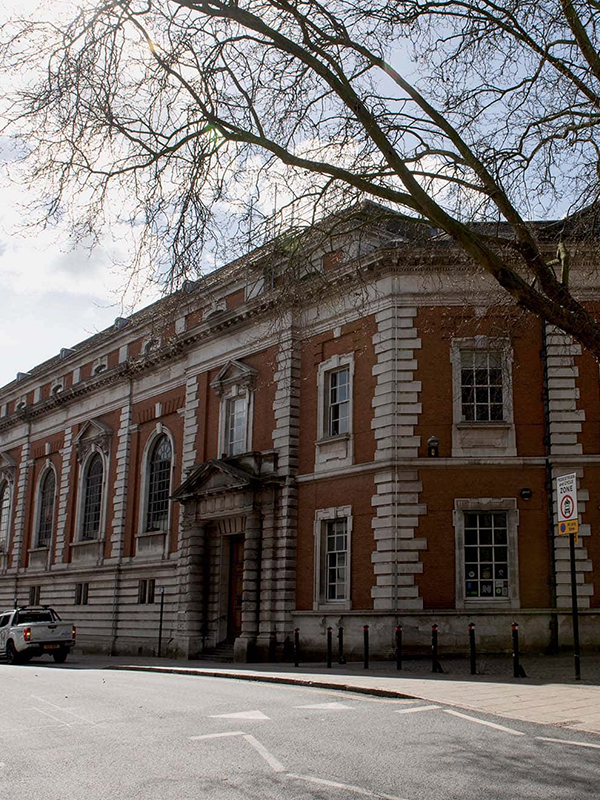 A historic brick building with large arched windows and a decorative entrance is surrounded by leafless trees. A parked vehicle is visible on the street in front, under a clear sky.