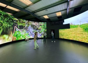 Three people stand in a circular room surrounded by large screens displaying nature scenes, including greenery and a cross on a hill. The ceiling is industrial with fluorescent lights, and the floor is smooth and dark.