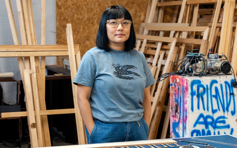 A person with straight black hair and glasses stands in an art studio. They wear a blue shirt with a bird graphic. Wooden frames and art materials are in the background.