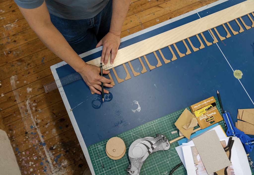 A person is working on a craft project at a blue table, using a compass to draw circles. The table has various tools, paper cutouts, a ruler, and a green cutting mat. The floor is wooden, and the person wears a gray shirt.