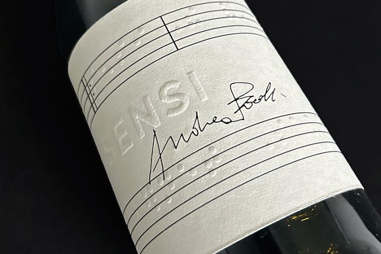 A close-up of a wine bottle with a white label featuring embossed braille and the name "Sensi" alongside a handwritten signature.