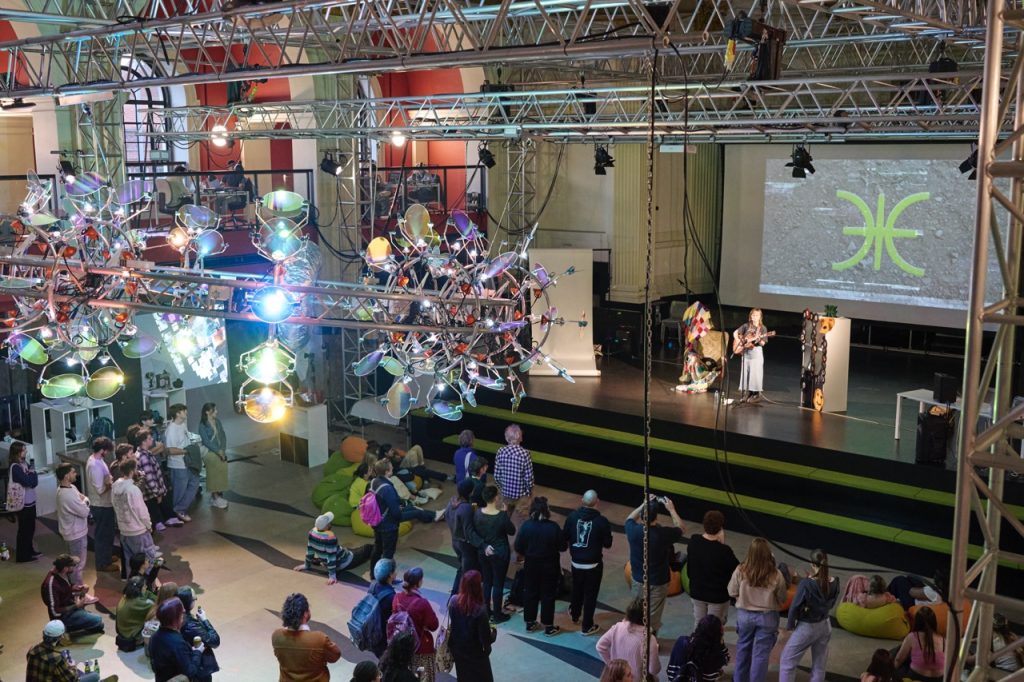 A large indoor event with people gathered around a stage watching a presentation. The stage features two speakers, and a screen displaying a symbol. Overhead, artistic light fixtures hang from the ceiling. Attendees sit and stand, observing the event.