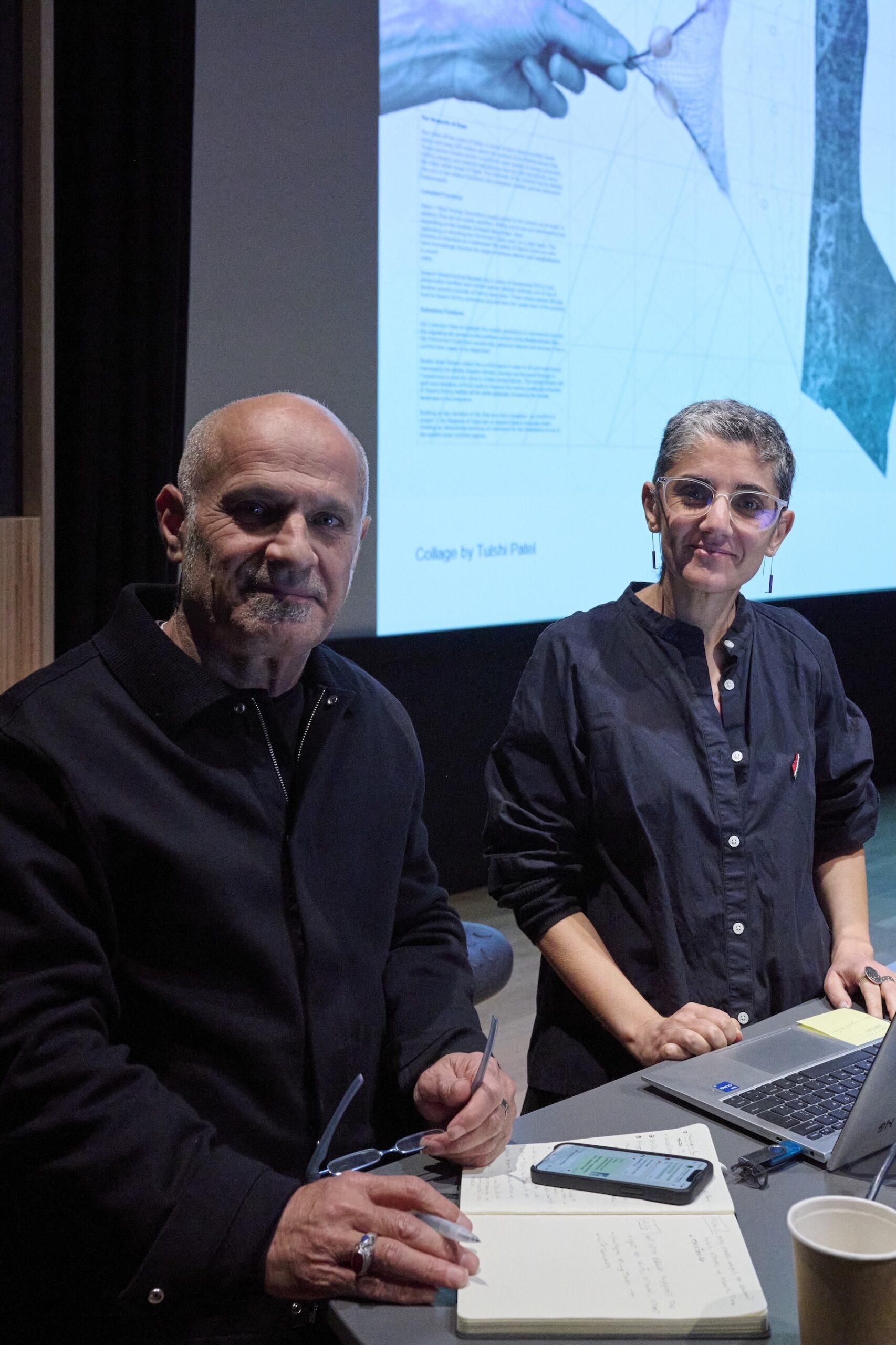 Two people stand at a table with notebooks, a phone, and a laptop, looking at the camera. Behind them, a large screen displays a collage and text, setting a thoughtful mood for their discussion on architectural humanities in the dimly lit room.