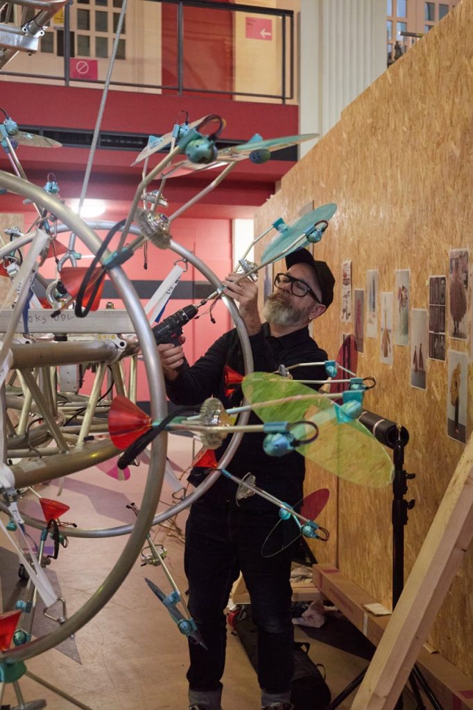 A person wearing a cap and glasses works on a large, abstract art installation featuring various colourful objects and metal rods. They are using tools and standing in a cluttered studio environment with art on the walls.