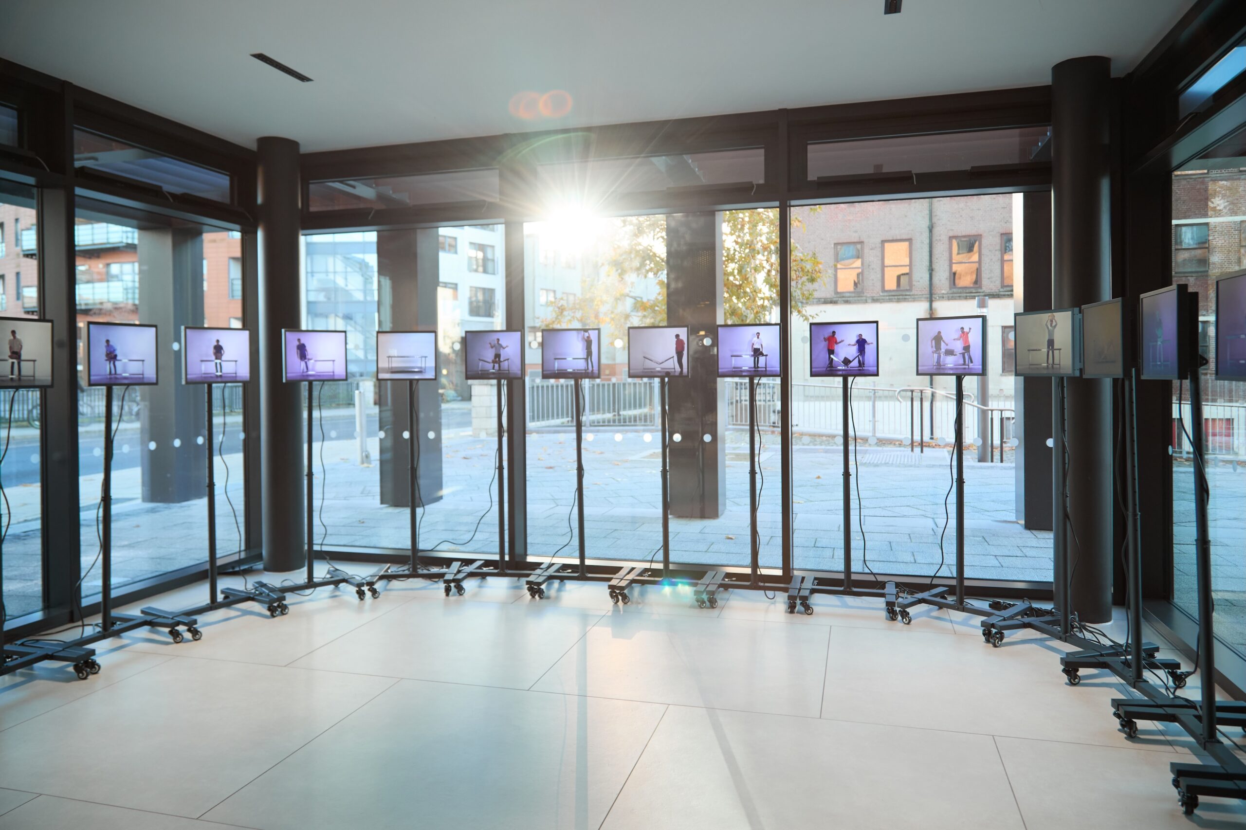 A sunlit room with large windows displays multiple screens on stands, arranged in a semi-circle, each showing images of a person in different poses—an engaging exploration of architectural humanities. Urban buildings are visible outside.