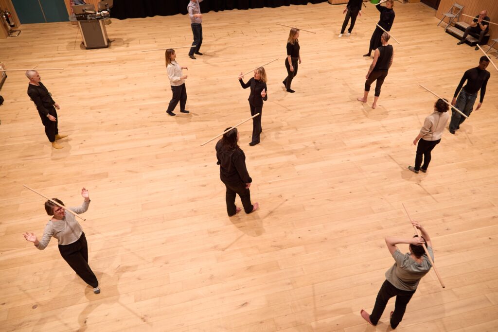 A group of people stand spaced apart on a wooden floor, each holding a wooden stick, participating in what appears to be a movement or martial arts class within an environment that reflects elements of architectural humanities.