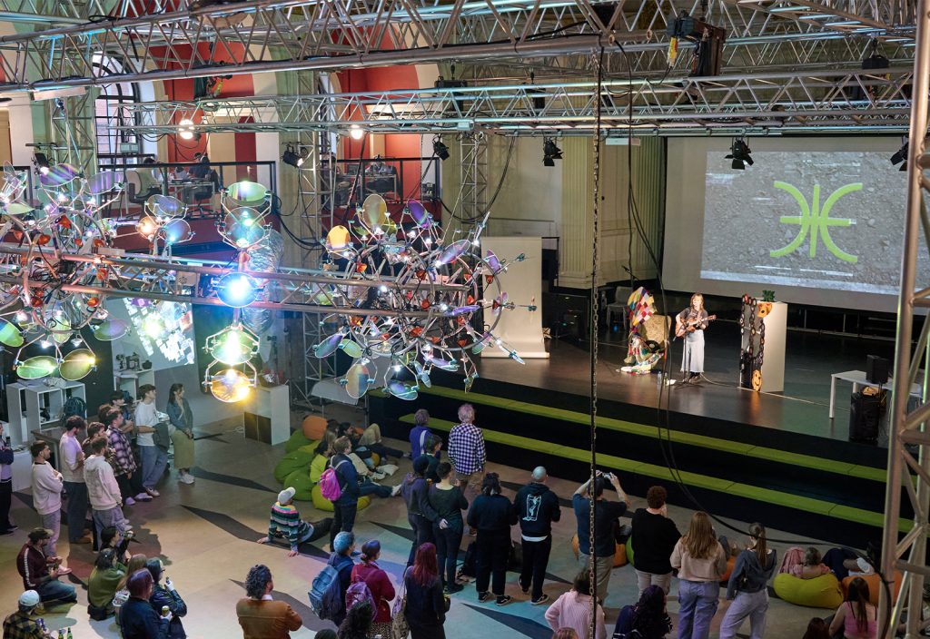 A crowd gathers in a spacious hall decorated with metallic sculptures and colourful lighting. They watch a presentation on stage, featuring a large screen displaying a symbol. The room has an industrial ceiling with exposed beams.