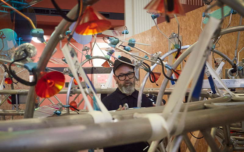 A person wearing glasses and a black cap is working on a complex metal structure adorned with colourful discs and wires, set indoors against a backdrop of wooden panels.