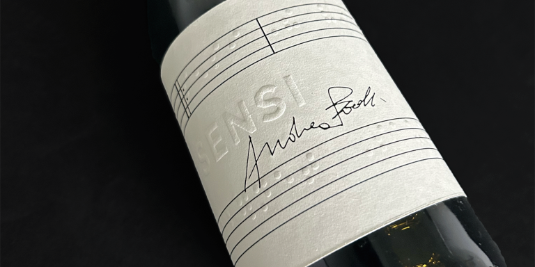 A close-up of a wine bottle with a white label featuring embossed braille and the name "Sensi" alongside a handwritten signature.