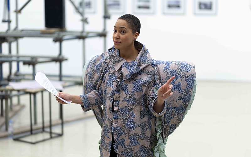 A person stands in a room holding papers, wearing a patterned, sculptural jacket with rounded shoulders. They appear to be speaking or gesturing, and there are blurred objects and framed photos in the background.