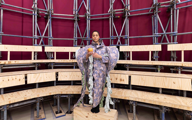 A person stands on a wooden platform in front of tiered seating made of metal and wood. They wear an elaborate, patterned outfit with large, wing-like extensions and hold an orange ball. The background consists of maroon fabric.