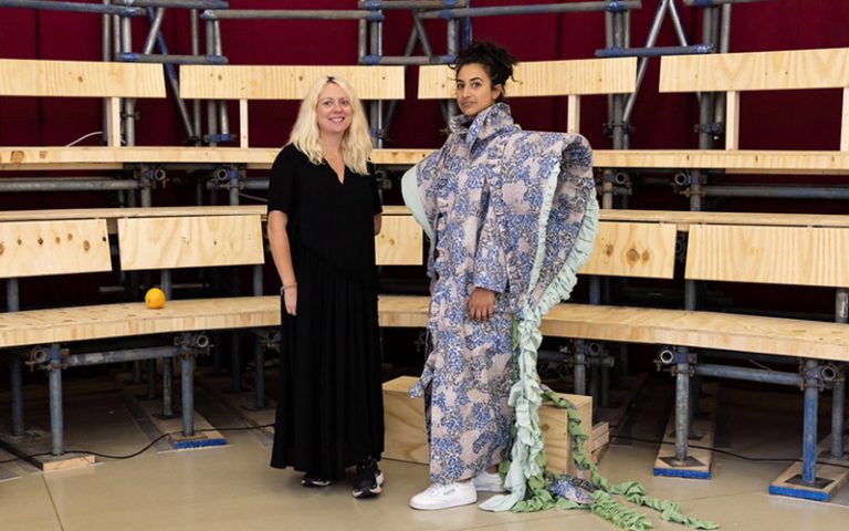 Two people stand in front of wooden bleachers. One wears a black dress, and the other wears a unique, patterned outfit with ruffled details and white sneakers. An orange sits on the ground nearby.