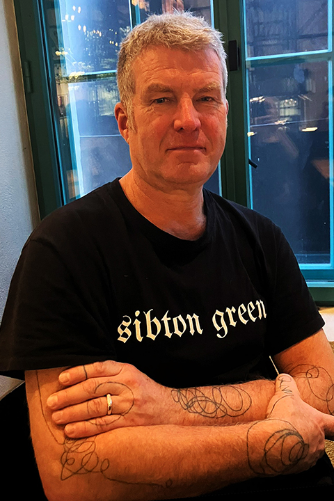 A person with short gray hair and tattoos on their arms sits with folded arms in front of a window. They are wearing a black t-shirt with the text "sibton green." The background shows a softly lit indoor setting.