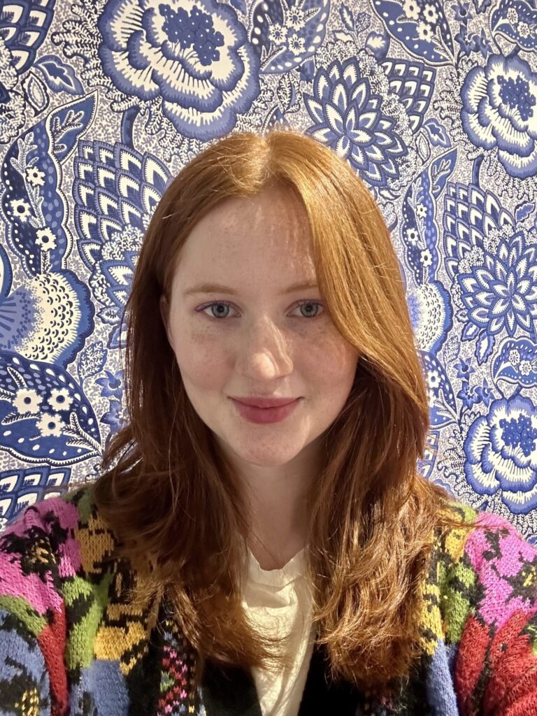 A person with long red hair and a patterned sweater stands in front of a blue and white floral wallpaper. They are smiling at the camera.