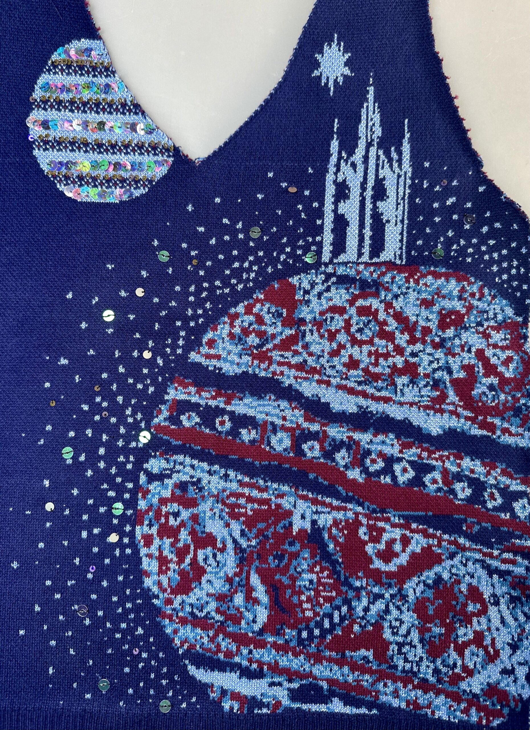 A detailed fabric design featuring a large planet with intricate red and pale blue patterns against a dark blue background. A smaller, colourful planet with multicolored sequins sits in the top left corner. Stars are scattered in the background.