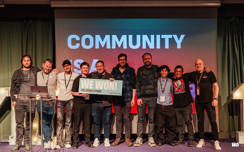 A group of nine people smiling and standing on a stage. Two individuals in the center hold a sign that says "WE WON! THE JUDGE'S PRIZE." The background features the word "COMMUNITY." They appear to be at an event celebration.