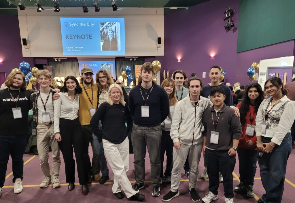 A group of people posing for a photo in front of a stage set up for a keynote speech. The background features a projection screen with the event name "Sync the City" and decorations with blue balloons.