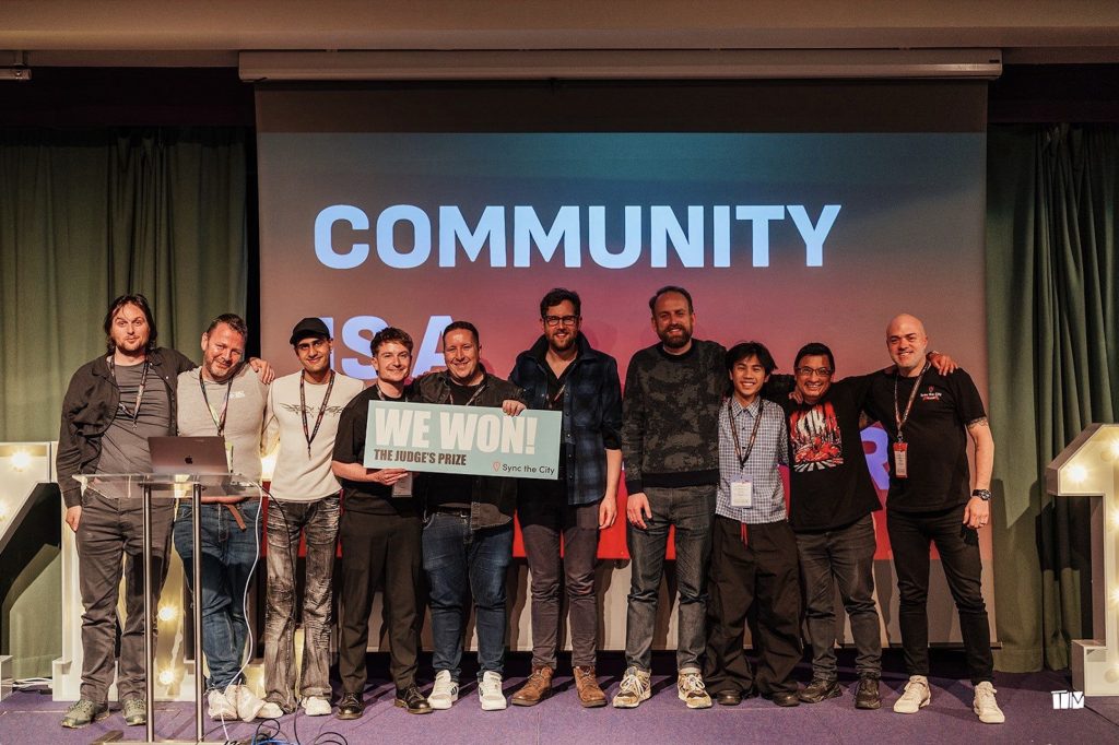 A group of people standing on a stage, smiling and holding a sign that reads "WE WON! THE JUDGE'S PRIZE." The background shows the word "COMMUNITY" in large letters. They appear to be celebrating their achievement.