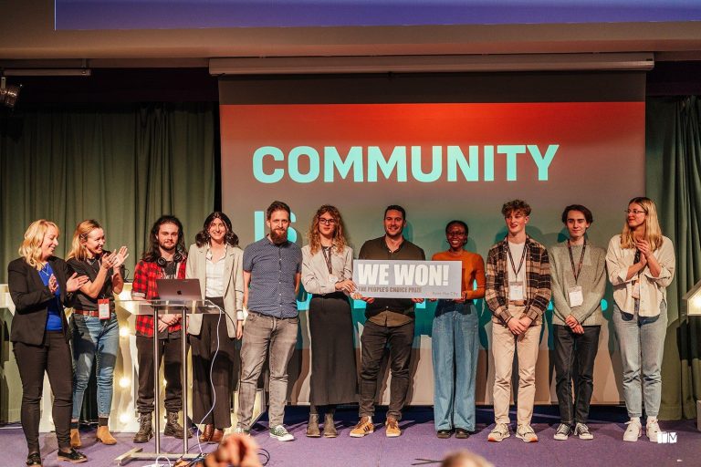 A group of people stands on a stage, some holding a sign that reads "WE WON!" in front of a large screen with the word "COMMUNITY" displayed. They appear to be celebrating an achievement.