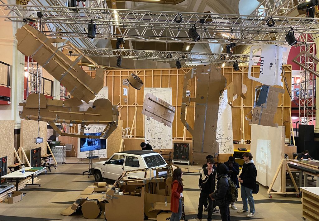 A large indoor art installation features various cardboard figures and structures suspended from a metal frame. A white car is visible beneath them. Several people are observing the exhibit. The background has wooden panels and lighting equipment.