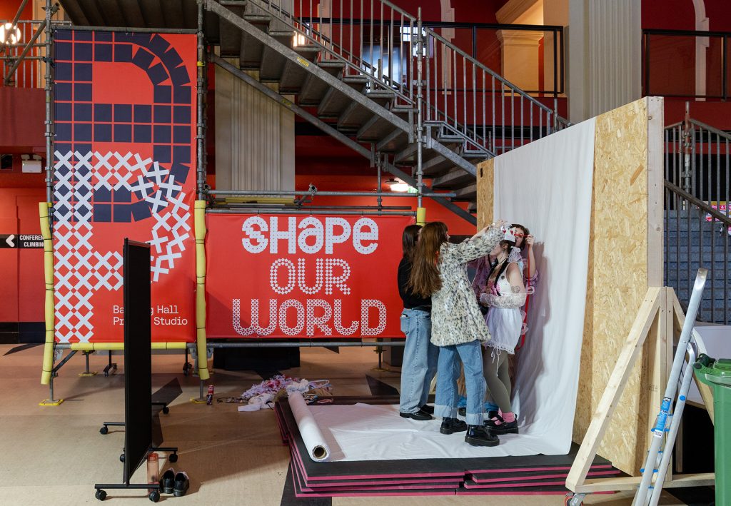 Two people adjust clothing on a model in a makeshift studio with a large "SHAPE OUR WORLD" banner in the background. The setting appears creative, with stairs and colourful walls.