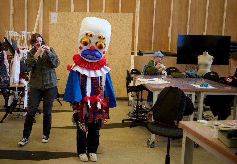 A person wearing a colorful, elaborate costume with a large, white headdress featuring multiple eyes and facial features stands in a workshop. Another person is taking a photo of them, and others are working at tables in the background.