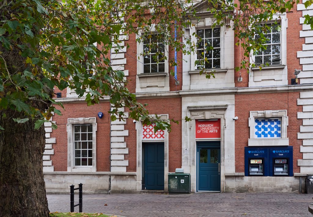 A brick building with the "University of the Arts" sign above a green door. Two ATMs are on the right. Trees with green leaves partially frame the scene. The area is cobbled and features a small black barrier to the left.