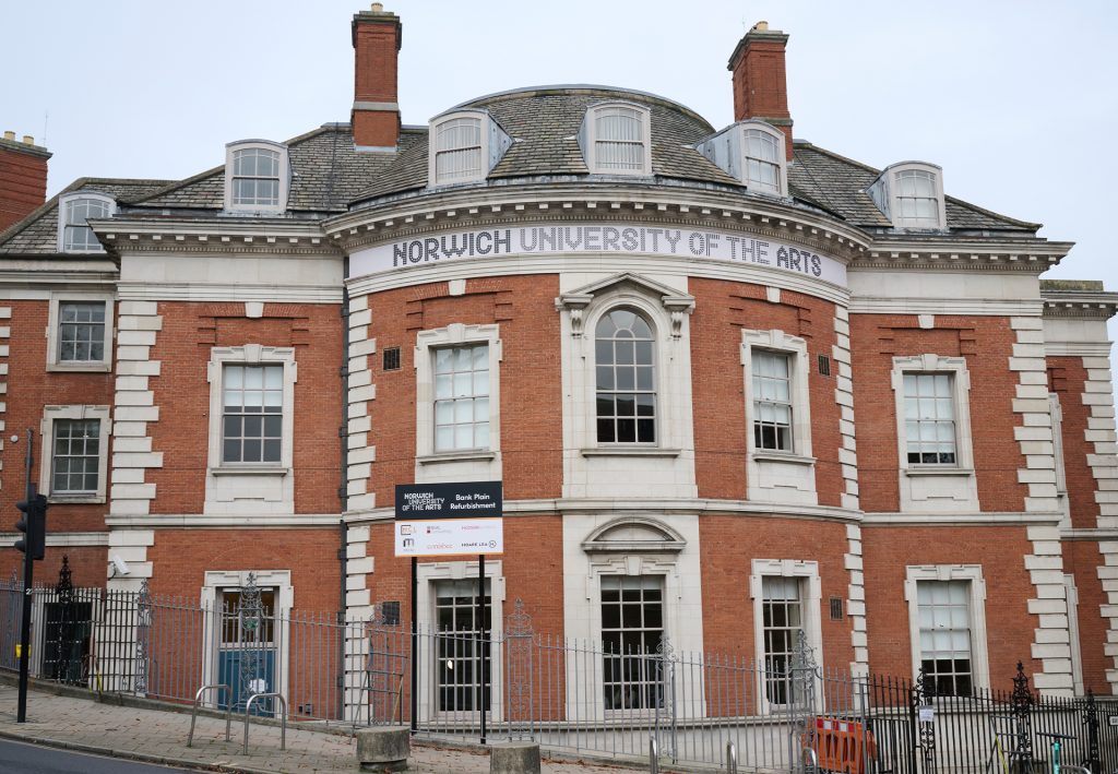 A red-brick building with white accents and a domed roof, featuring "Norwich University of the Arts" on a banner. The facade has large windows and a black sign near the entrance. The building is surrounded by a metal fence.