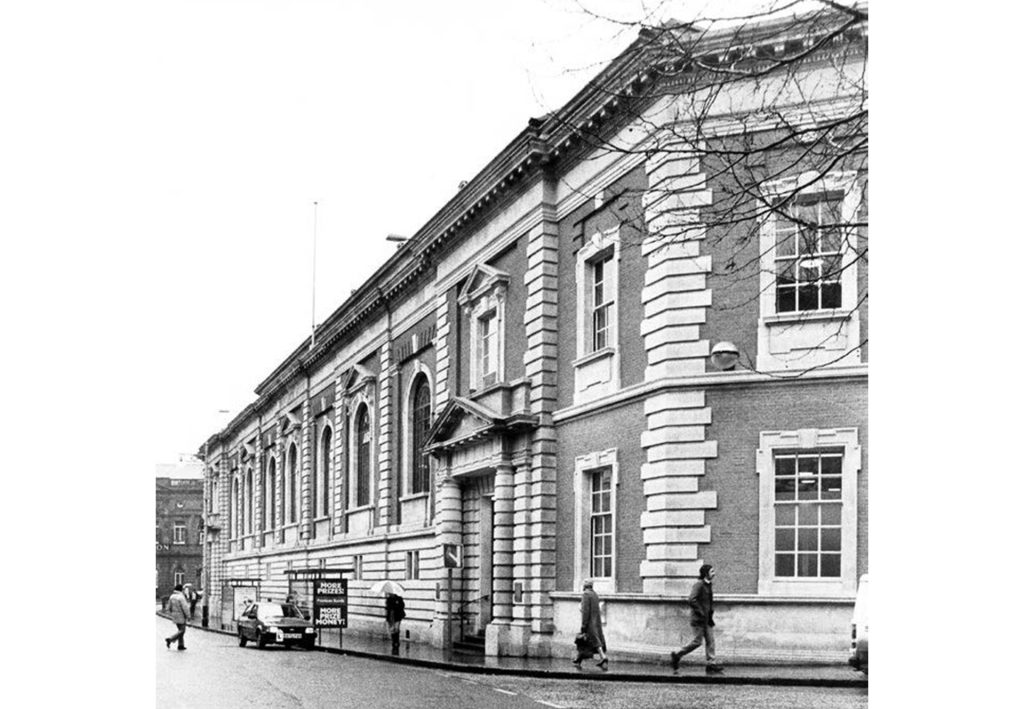 Black and white image of a historic building with ornate architecture, featuring large arched windows and detailed stonework. A few people walk along the sidewalk near a parked car in front of the building.