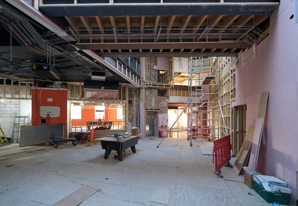 A large indoor construction site featuring unfinished walls and exposed beams. Scaffolding and ladders are set up, and there's a table saw and various construction materials scattered on the floor.