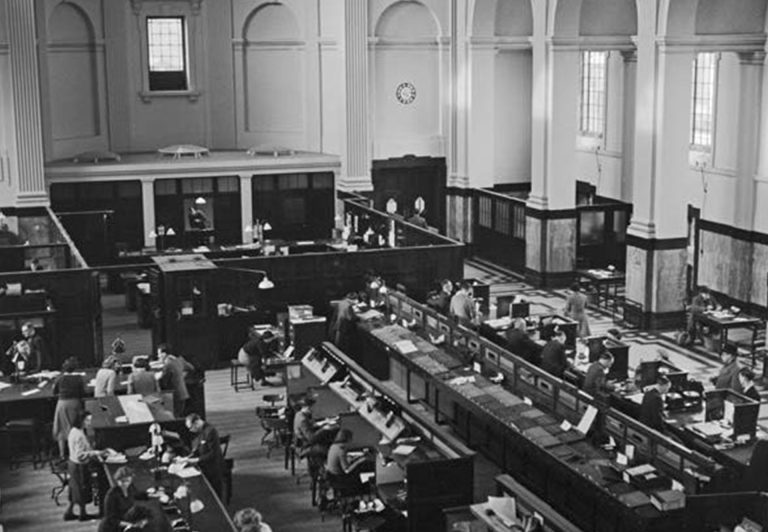 A black and white image of a vintage bank interior, featuring high ceilings, large windows, and busy counters. People are seen conducting transactions and working at desks. The setting has an early 20th-century architectural style with classical details.