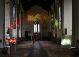 A dimly lit church interior features colourful, abstract light projections on the walls and ceiling. The space includes rows of benches, stained glass windows, and a display case with illuminated art pieces on either side of the aisle.