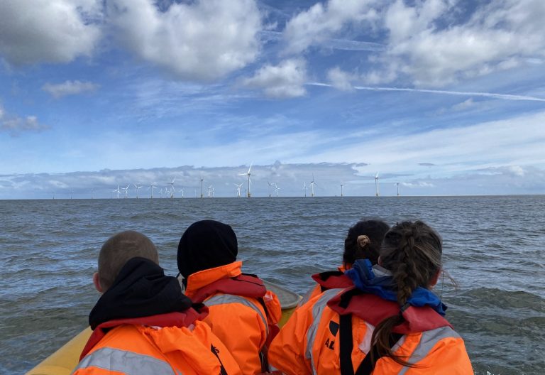 Three people in orange life jackets sit in a boat, facing away and looking at an offshore wind farm on a cloudy day.