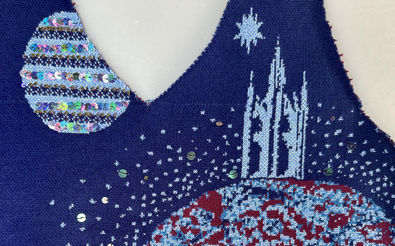 A section of a knitted artwork features a celestial theme with a round, striped planet on the left and a tall, intricate structure resembling a castle on the right. The background is dark blue, adorned with scattered sequin-like stars and a prominent white star.