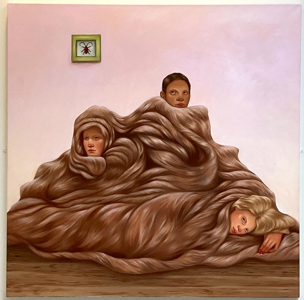 Surreal artwork shows three human faces partially enveloped in large, flowing brown hair, covering their bodies like a blanket. A small framed image of a beetle hangs on the plain pink wall in the background.