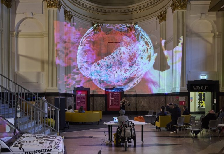 A large, abstract, colourful digital projection fills the wall of an ornate interior space with columns and gold accents. People sit on yellow couches and chairs, observing the vibrant display.