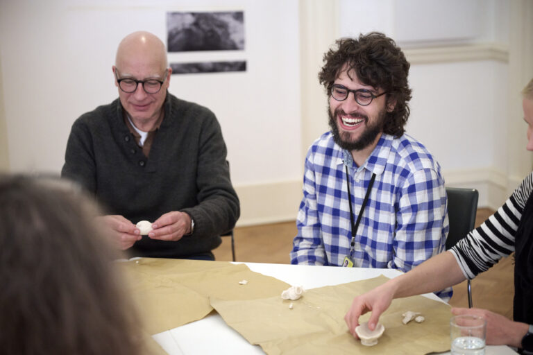 People sat around a table working on a paper project. One person in glasses and with curly hair and a beard is laughing and smiling