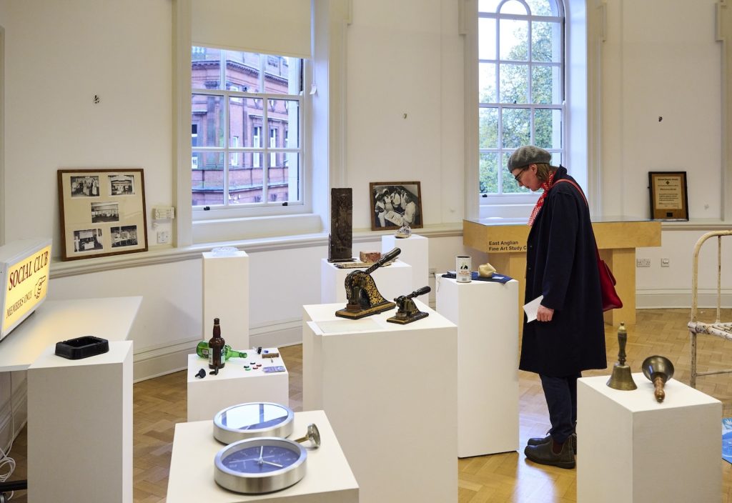 A person wearing a cap and coat observes various objects on white pedestals in an art gallery. Items include vintage photos, a bottle, mechanical parts, a clock, and a bell. Large windows in the background provide natural light.