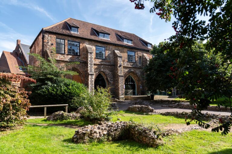 A landscape photo of East Garth, Norwich University of the Arts's photography building. The building is old and has flint features. In front of the building are scattered remains of flint wall amongst green grass and trees.