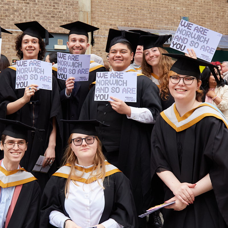 A group of students are stood together wearing their graduation robes and mortarboards. They are all holding a lilac card that says 'We are Norwich and so are you'