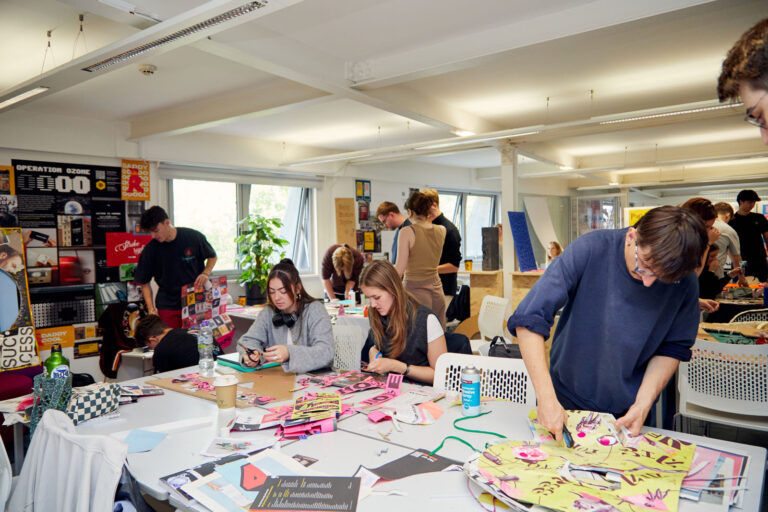 In the busy graphics studio, students are sat and stood around tables working together, cutting up vibrant sheets of paper with typography on