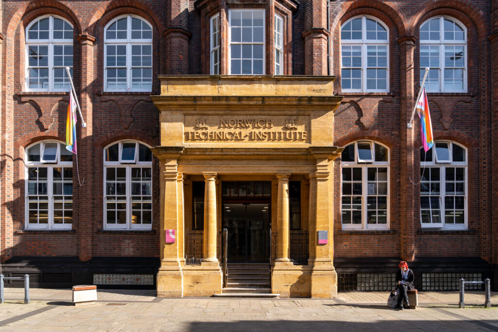 A photo of Norwich University of the Arts' St Georges Building. Either side of the entry, are two flagpoles with the LGBTQIA+ flag on.