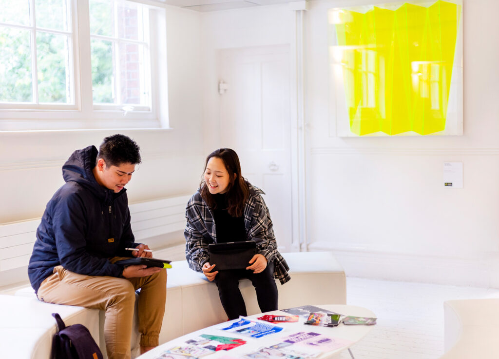 Two students are sat on a white bench, in front of a white wall, talking and looking at an iPad. On the white coffee table in front of them are a variety of colourful flyers and leaflets