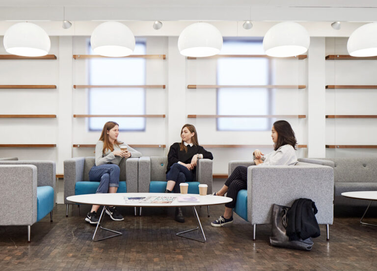 Three students are sat in individual grey armchairs, with a white oval coffee table in front of them. The students are talking to each other whilst having a coffee.