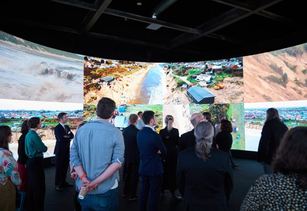 A group of people stands inside a room with large, curved screens displaying aerial and close-up images of a coastal area, showing erosion and urban settings. The audience appears engaged in observing the visuals.