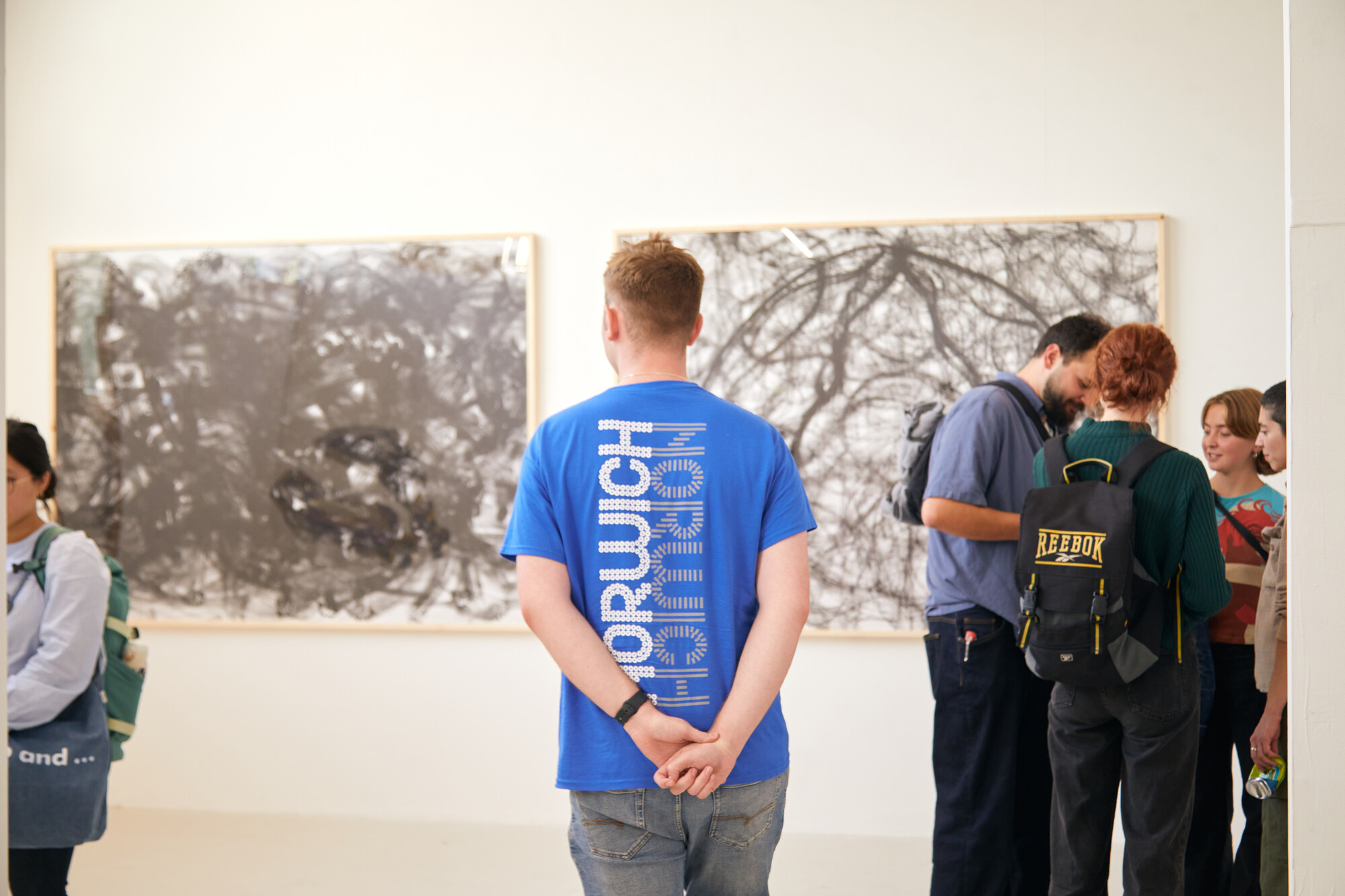 Ambassador standing in Grad Fest during degree shows, his back is facing the camera, blue Norwich Ambassador top. Visitors on either side of him.