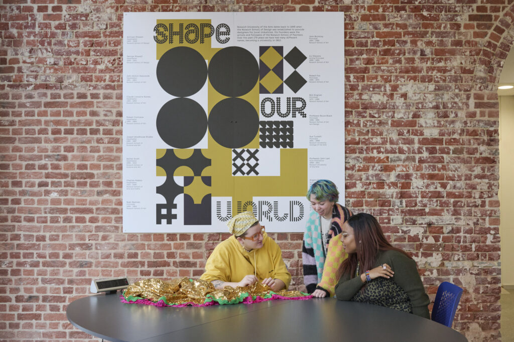 Three students sitting around a table with banner behind 'Shape our World'