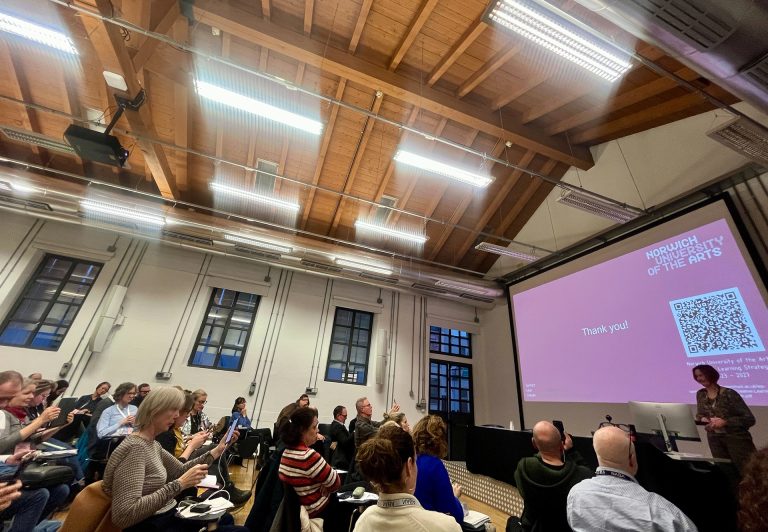 A presenter from Norwich University of the Arts addressing an audience in Milan. The audience is seated, some are clapping. A screen displays a "Thank you!" slide with a QR code. The room has a high wooden ceiling and large windows.