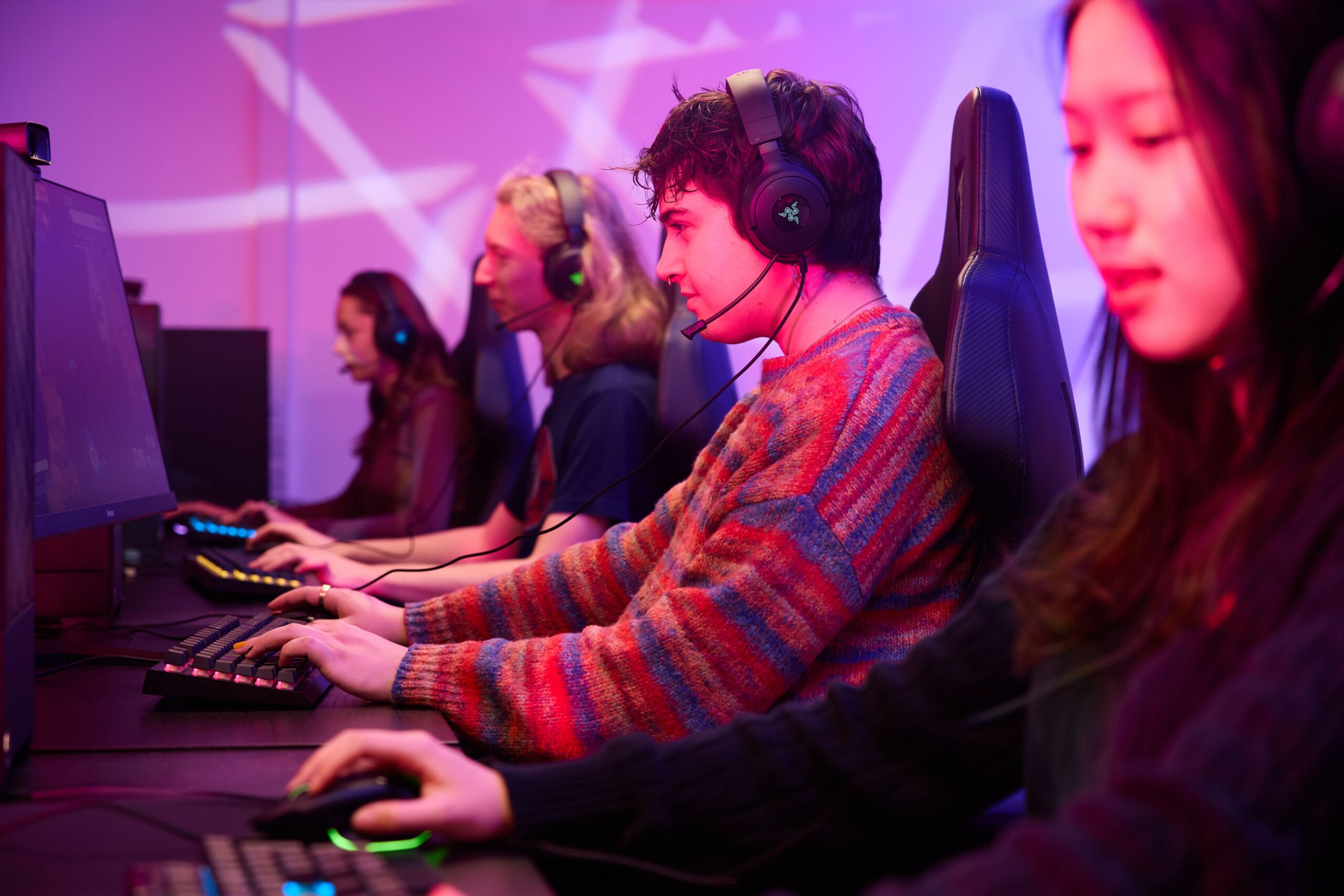 Four young people wearing headsets sit in a row at computers, intensely focused on gaming in a room lit with pink and purple lights.